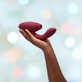 A person holds a Bordeaux Womanizer Duo 2 Rabbit Air Pulse Vibrator, showcasing Pleasure Air Technology, against a softly blurred background of pastel bokeh lights.